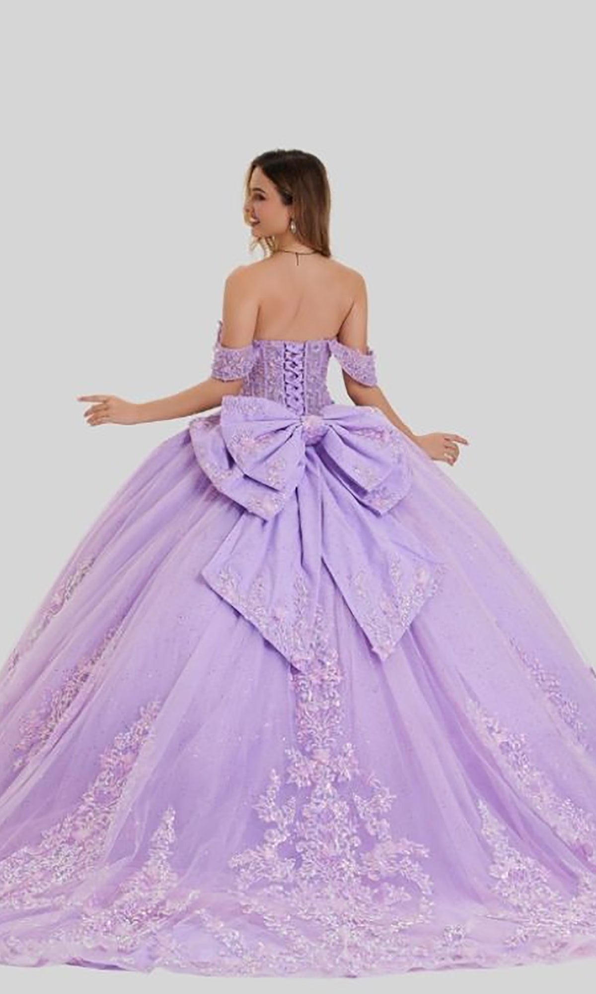 A woman wearing the Quinceanera Dress 26408 By Stella Couture—a stunning off-the-shoulder ball gown in lavender with delicate floral embroidery and a large bow on the back—stands against a plain light background.